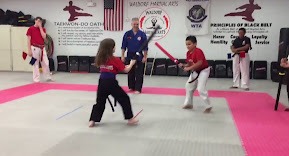 Two martial arts students practice controlled sparring drills while wearing protective equipment on training mats.