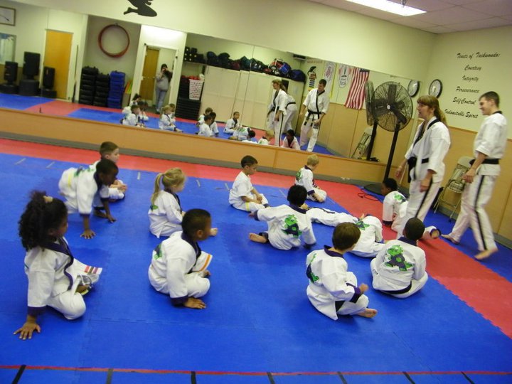 Martial arts students seated on mats participate in a structured class activity within the training space.