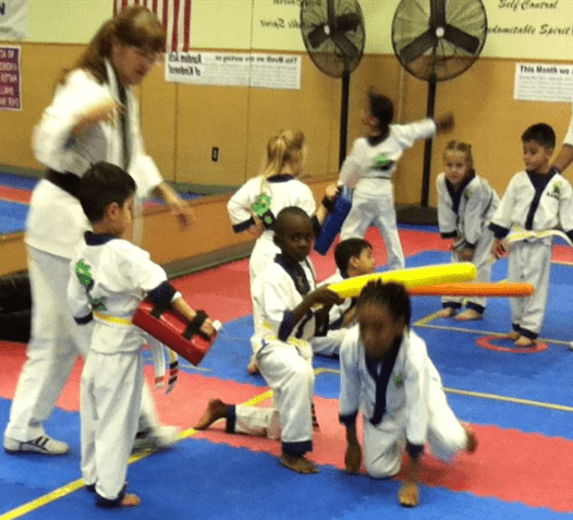 Children in martial arts uniforms practice techniques, while an instructor guides them. The vibrant classroom features training tools and encourages teamwork.