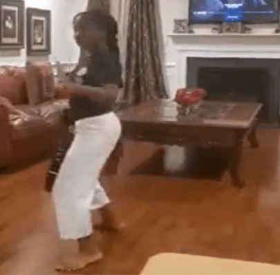 A young girl dances barefoot in a living room, wearing a black top and white pants, with a coffee table and a television in the background.