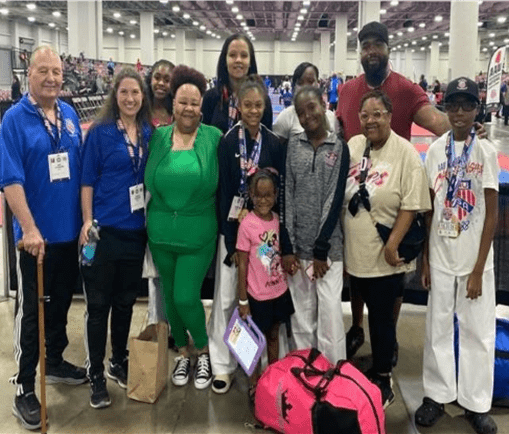 A diverse group of ten martial artists and coaches poses together at a tournament venue, showcasing team spirit and camaraderie.