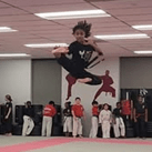 A young martial artist performs a high jump kick in a training dojo, showcasing skill and focus, with classmates observing in the background.