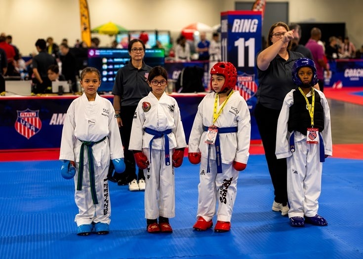 Four young martial artists in doboks stand ready at a competition, with one adult overseeing. They're poised for a match at Ring 11.