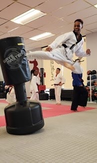 A martial artist performs a high kick against a training dummy in a dojo, demonstrating skill and technique amid fellow practitioners.