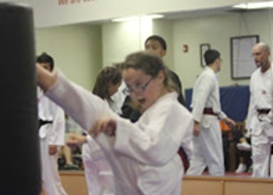 A young girl in a karate uniform performs a high kick against a padded target, surrounded by fellow practitioners in a martial arts class.