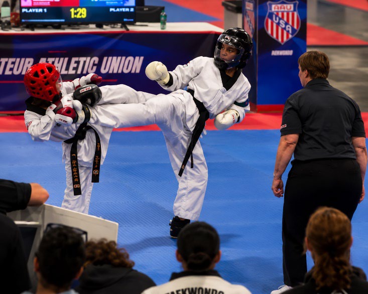 Two athletes spar in a taekwondo competition, with one executing a high kick while the referee observes. The scoreboard shows 1:29 remaining.