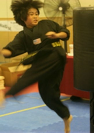 Young martial artist in black uniform executing a high kick towards a training bag, showcasing agility and skill in a dojo setting.
