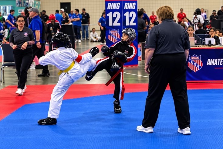 Young martial artists compete in a Taekwondo match, showcasing a kick. Officials and spectators observe closely in a tournament setting.