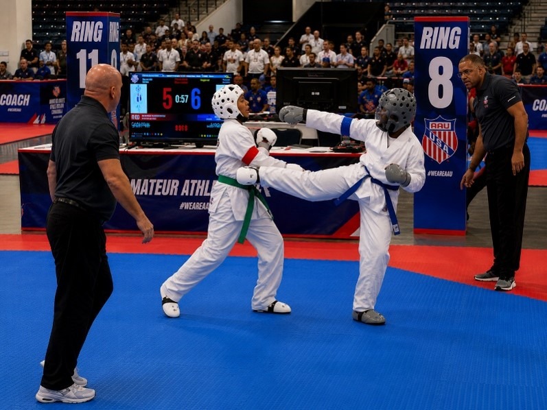 Two athletes spar in a karate match, one delivering a high kick while the other blocks. Officials and a scoreboard are visible, highlighting the competition.