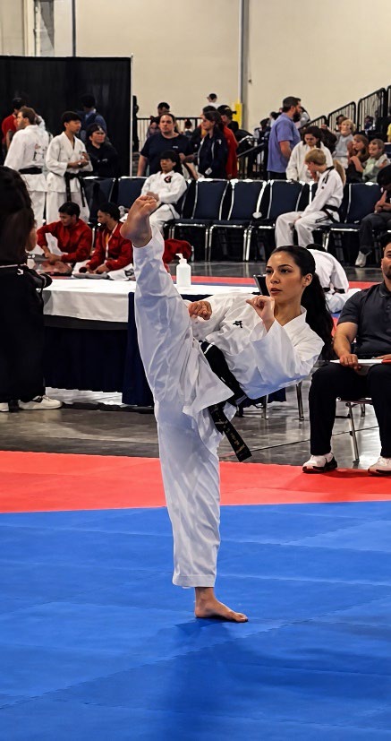 A black-belt female martial artist performs a high kick in a competition, surrounded by judges and spectators, showcasing skill and precision.