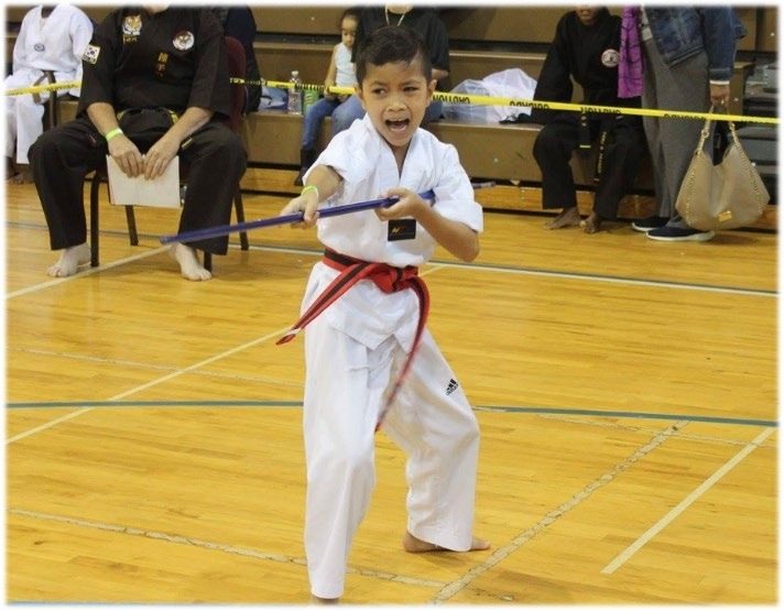 A young martial artist in a white uniform and red belt performs with a blue weapon during a tournament, showcasing skill and enthusiasm.