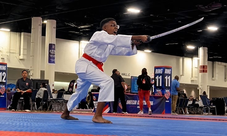 A young martial artist in a white gi with a red belt performs a kata with a sword, showcasing skill and focus during a competition.